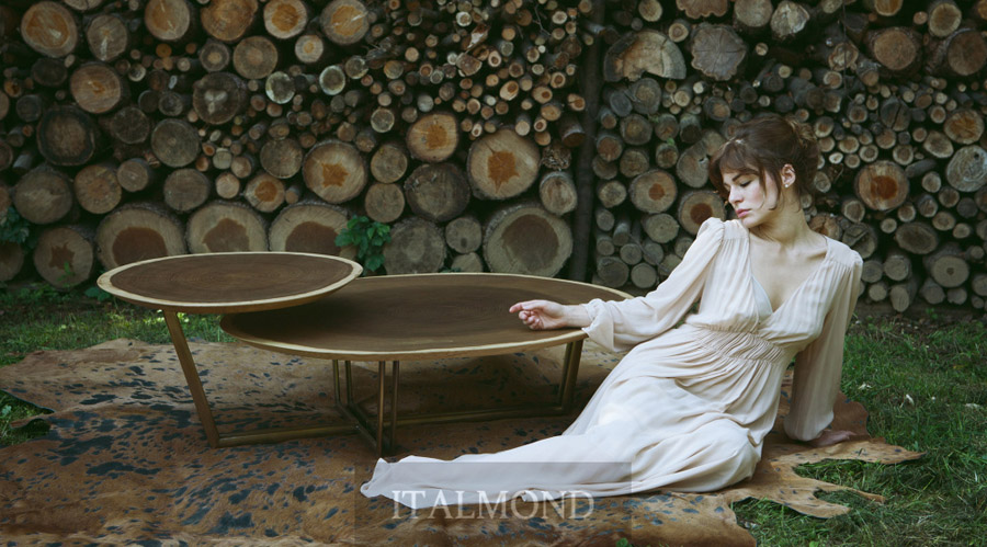 Женщина в белом платье сидит у деревянных столиков на фоне дровяника